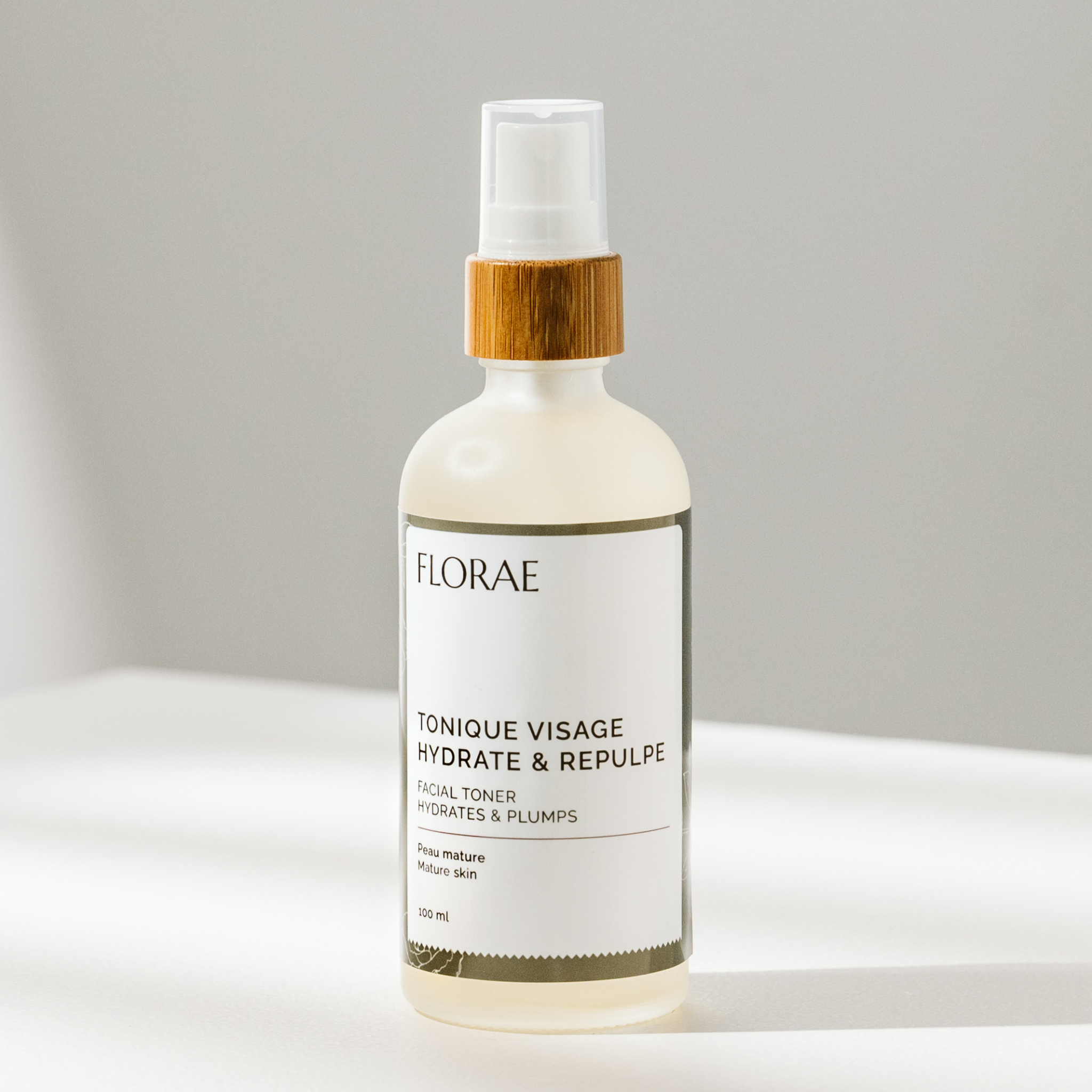 onique pour le visage de la gamme Florae cosmétiques. Présenté dans une bouteille de verre avec un bouchon de bambou vaporisateur, sur un fond blanc.