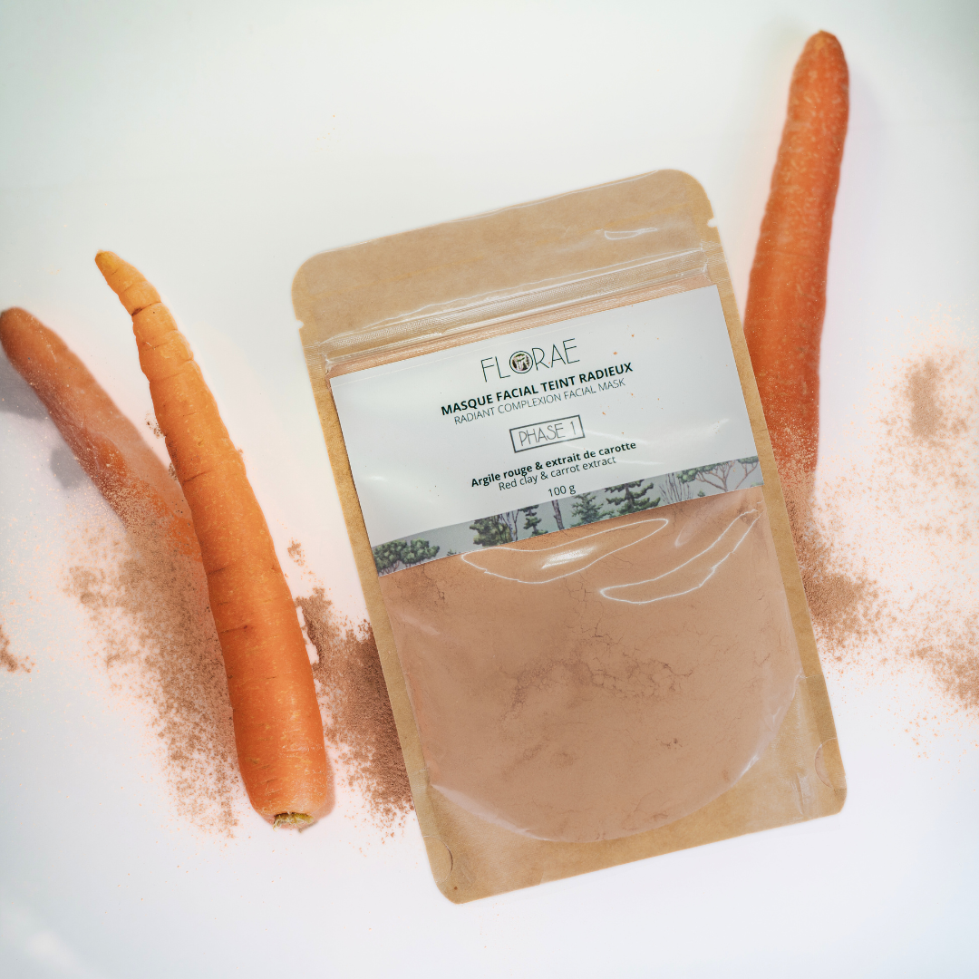 Sachet en paier kraft avec une fenêtre transparente en avant. On y voit la poudre orange. Présentation sur un fond blanc avec des carottes et de la poudre orangée dispersée. Actifs pour masque facial.